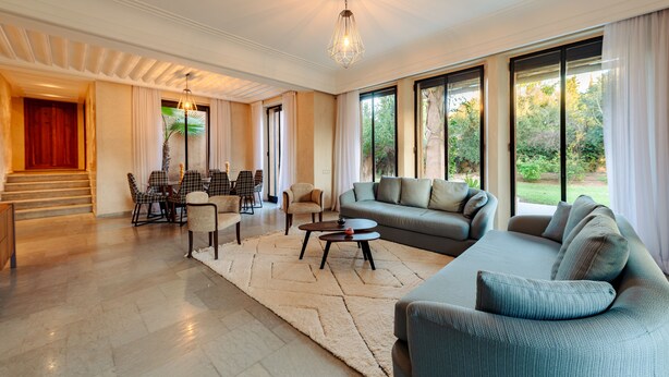A spacious living room with a large sofa, a coffee table, and a dining area with chairs. There is a staircase leading to another level, and large windows offering a view of the outdoors. - Villa de luxe Sahara  (Marrakech)
