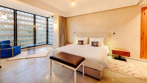 A spacious bedroom with a large bed, a bench, a small table, and a view through a window with a decorative screen. - Villa de luxe Sahara  (Marrakech)