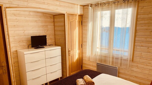 Ein Zimmer mit Holzwandverkleidung, einem Bett, einer weißen Kommode, einem Fernseher und einem Fenster mit durchsichtigen Vorhängen.