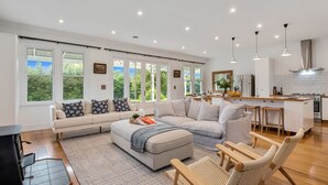 A modern living room with a sofa, ottoman, and wicker chairs, a kitchen with a bar area, and large windows with a view of greenery. - Botham's Beauty - Blenheim Holiday Home (Blenheim)