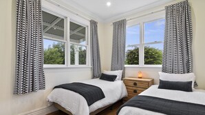 A bedroom with two beds, white bedding, black and white patterned curtains, a wooden nightstand with a lamp, and large windows with a view of greenery. - Botham's Beauty - Blenheim Holiday Home (Blenheim)