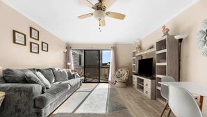 A living room with a grey sofa, a TV, a ceiling fan, and a dining area with chairs and a table. - Steps Away From The Ocean (North Myrtle Beach)
