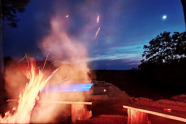 Ein Lagerfeuer mit fliegenden Funken, eine hölzerne Bank und ein Baum im Hintergrund.