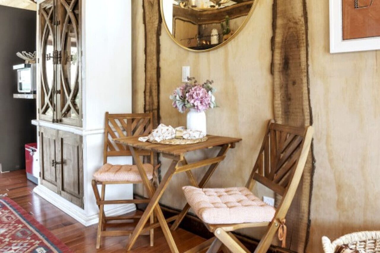 A dining area with a wooden table and chairs, a vase with flowers, and a mirror on the wall.