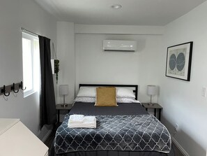 A neatly arranged hotel room with a bed, bedside tables, an air conditioning unit, a window with curtains, and a framed picture on the wall.