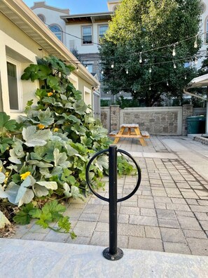 Un espacio al aire libre con suelo empedrado, una mesa de picnic de madera, un poste negro con un círculo en la parte superior y un árbol grande.