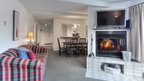 Un salon avec un canapé à carreaux, un téléviseur à écran plat, une cheminée et un coin repas comprenant une table et des chaises.