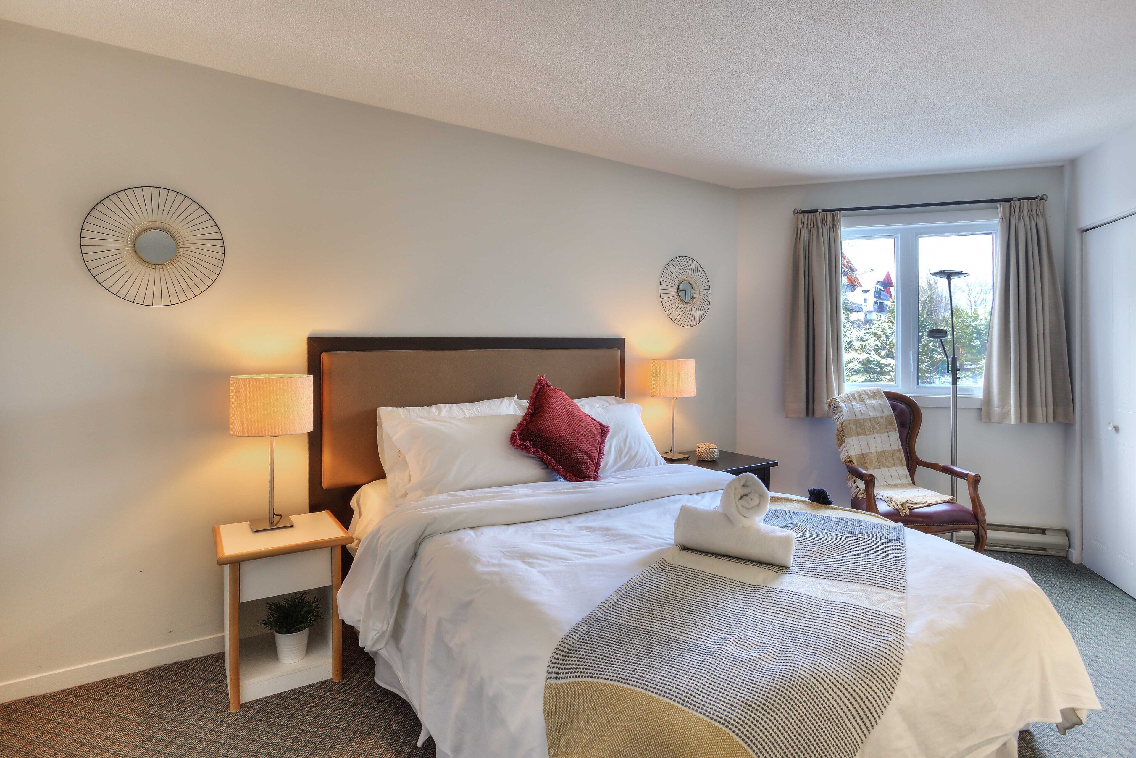A hotel room with a neatly made bed, two bedside lamps, a chair, and a view of the outdoors through a window.
