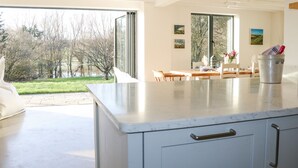 A modern kitchen with a large island, a dining area with a table and chairs, and a view of a garden and trees through large windows. - Meon Boscage (Southampton)