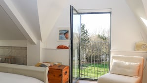 A bedroom with a bed, a wooden chest, a chair, and a balcony with a view of trees and grass. - Meon Boscage (Southampton)