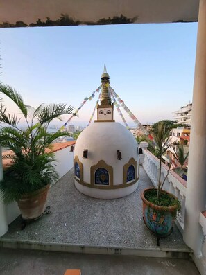 En lille, hvid, kuppelformet bygning med farverige vinduer og et flag på toppen, omgivet af plantekasser med blomster på en balkon med udsigt over bybilledet.