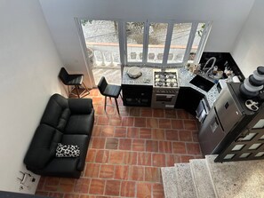 En kompakt opholdsstue med en sort lædersofa, et lille køkken med gasblus og et spiseområde med to stole.