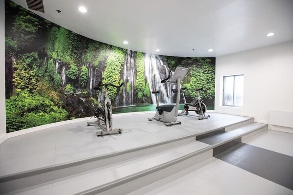 Stay active in our fitness room, featuring exercise bikes and a bench, with a scenic mural of a forest waterfall.
