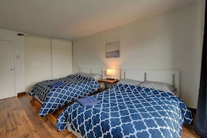 A bedroom with two beds, wooden flooring, a nightstand with a lamp, and a painting on the wall.