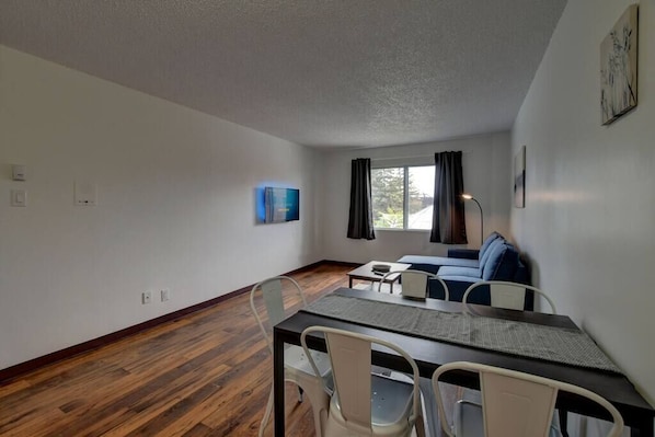 A room with a dining table, chairs, a sofa, a window with curtains, and a wall-mounted artwork.