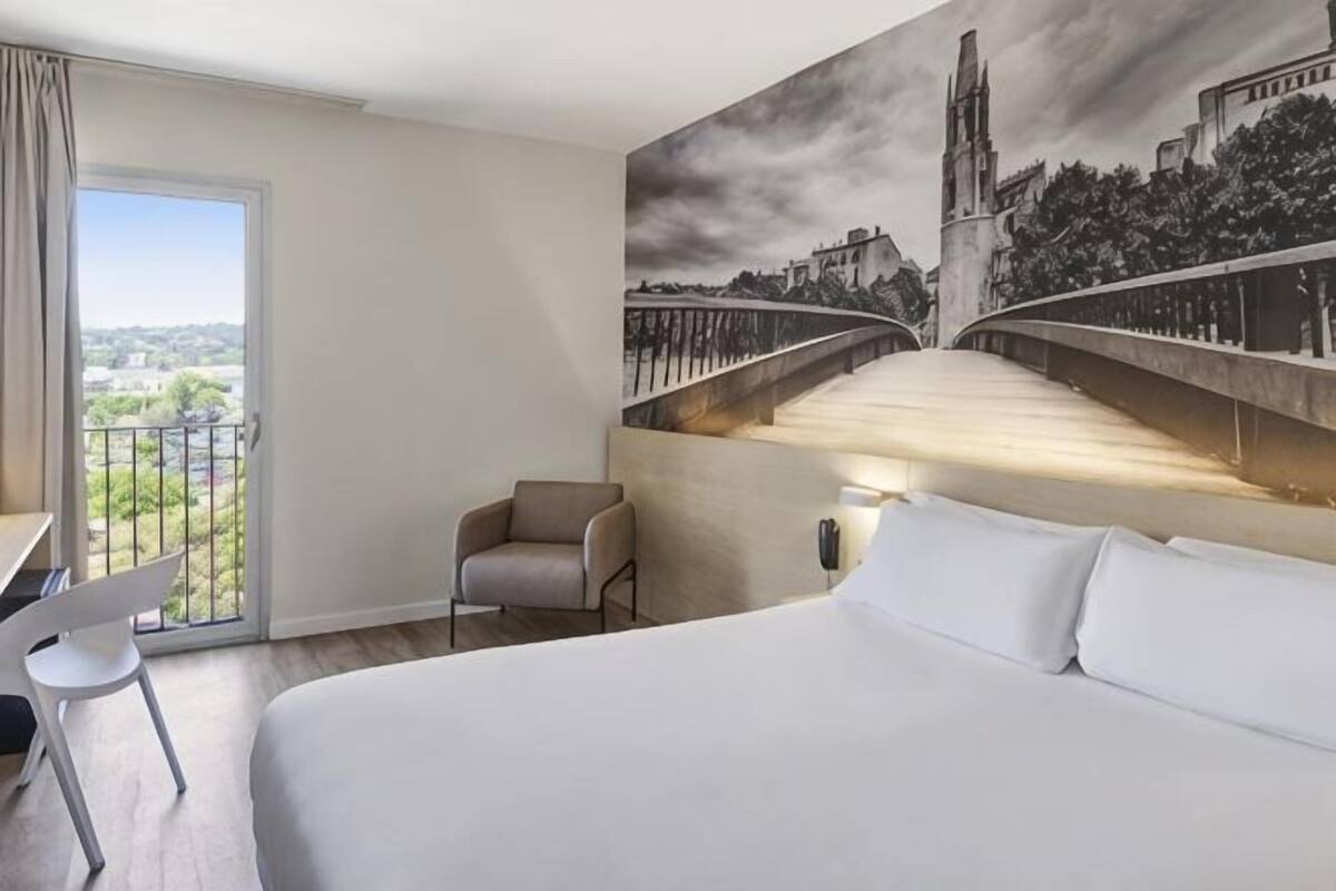 A hotel room with a bed, a chair, a desk, and a painting of a bridge.