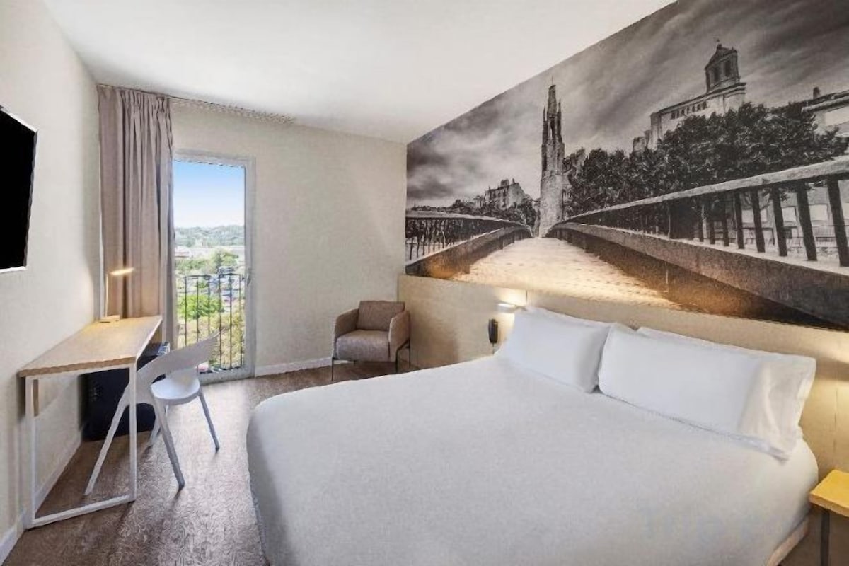 A hotel room with a bed, a desk, a chair, and a large window.