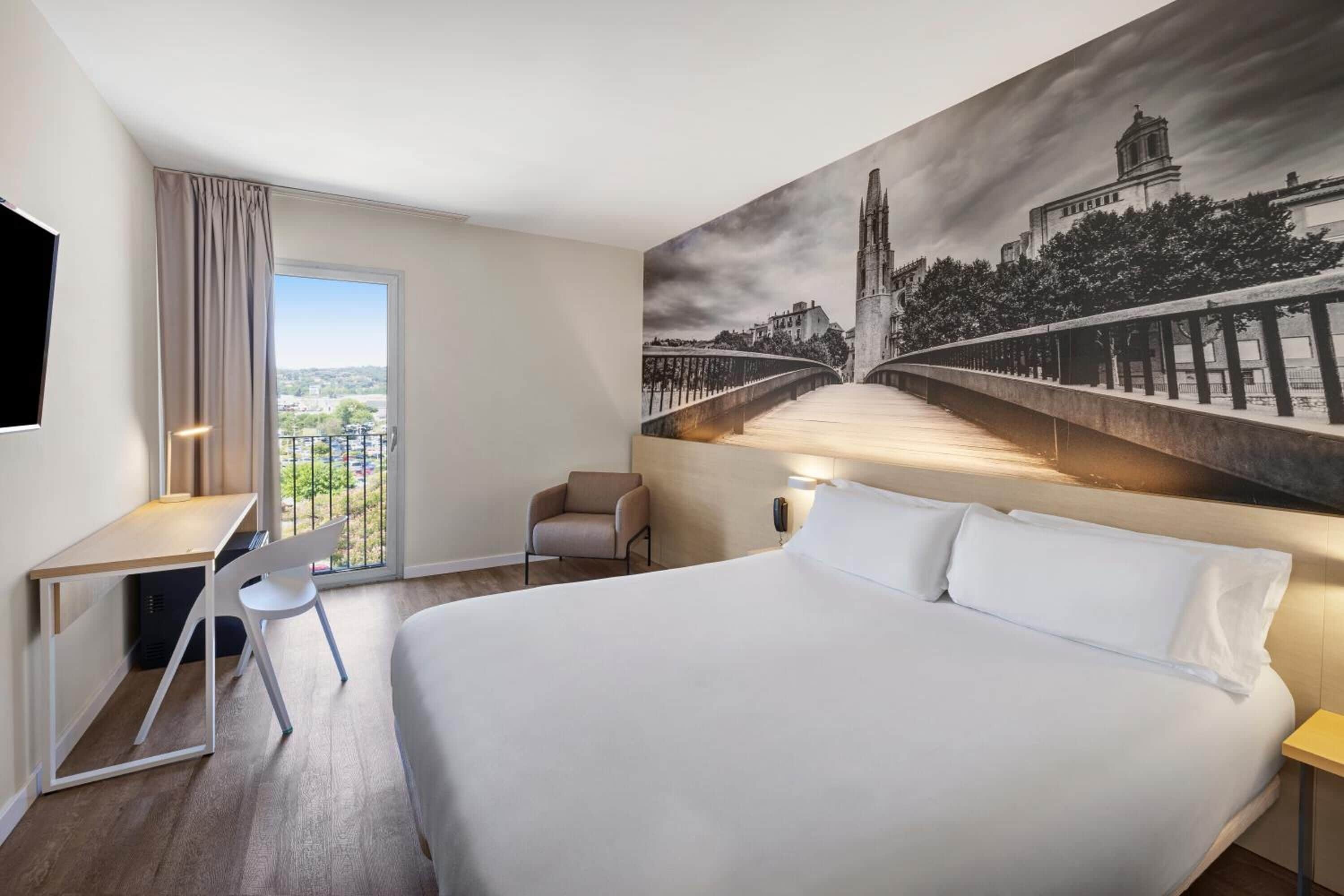 Une chambre d’hôtel avec un grand lit, un bureau, une chaise et une vue sur un pont et le paysage urbain, le tout visible à travers une grande fenêtre.