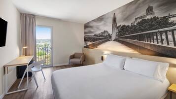 Une chambre d’hôtel avec un grand lit, un bureau, une chaise et une vue sur un pont et le paysage urbain, le tout visible à travers une grande fenêtre.
