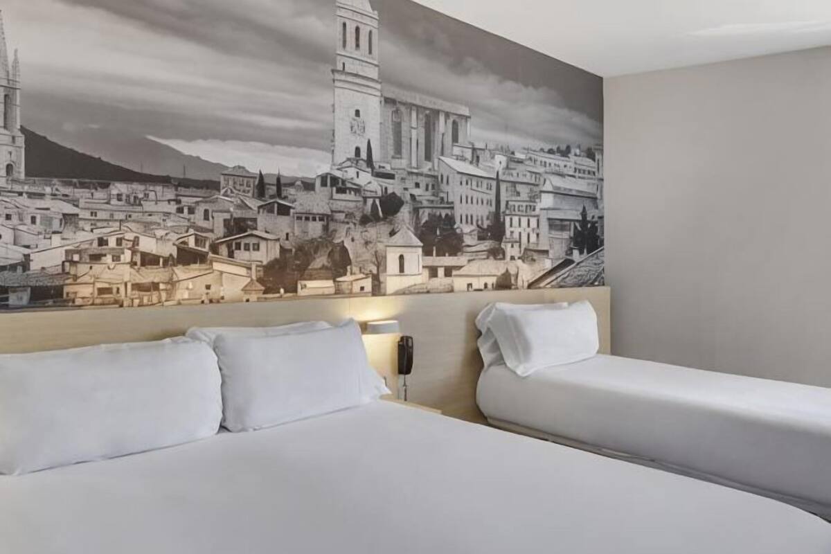 A hotel room with a large cityscape mural, two beds, and a bedside table.