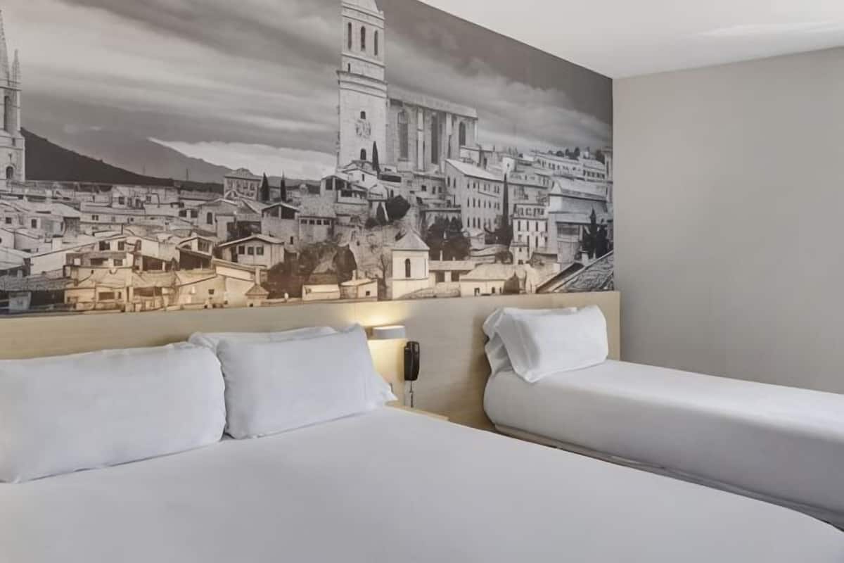 Habitación de hotel con un gran mural de paisaje urbano, dos camas y una mesita de noche.