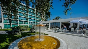 Una fuente en una cuenca circular, rodeada por una zona pavimentada con asientos exteriores. En el fondo, se observa un edificio moderno con varios pisos y grandes ventanales.