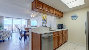 Una cocina moderna con armarios de madera, lavavajillas e isla grande. Hay una zona de comedor con mesa y sillas, y vistas de la ciudad a través de amplias ventanas.