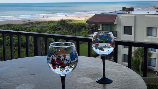 Dos copas de vino en un balcón con vistas a una playa y al océano, con un edificio en el fondo.