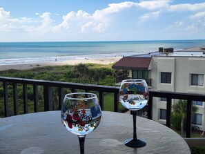 Two wine glasses on a balcony overlooking a beach with a view of the ocean and a building in the background.