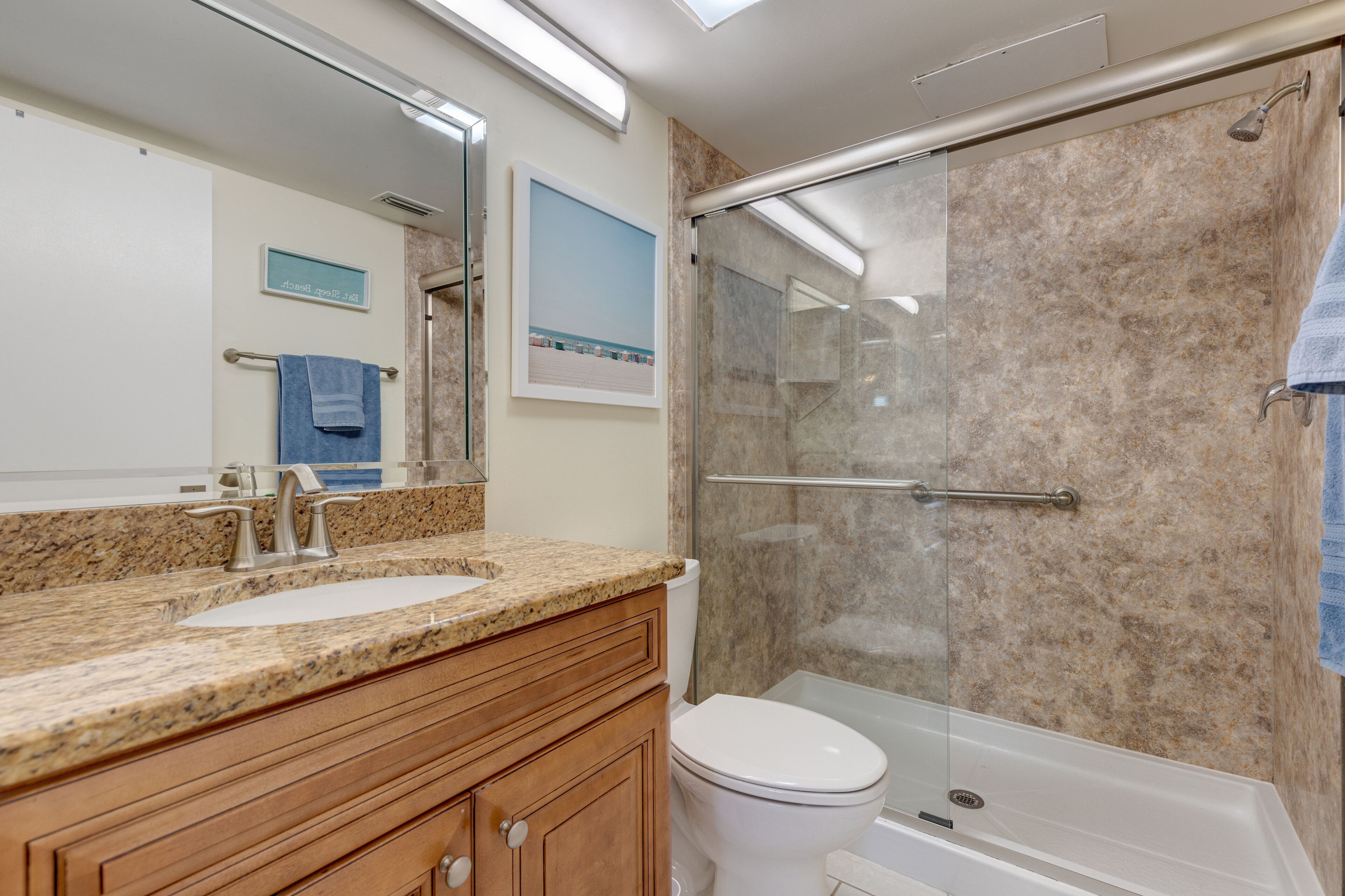 Un baño con encimera de granito, lavabo blanco, tocador de madera, inodoro blanco y cabina de ducha con mampara de vidrio.