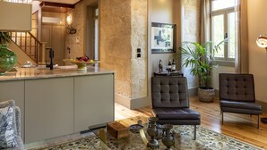 A spacious living room with a glass coffee table, two dark armchairs, a sofa, a kitchen area with a fruit bowl, and a staircase with exposed wooden beams.