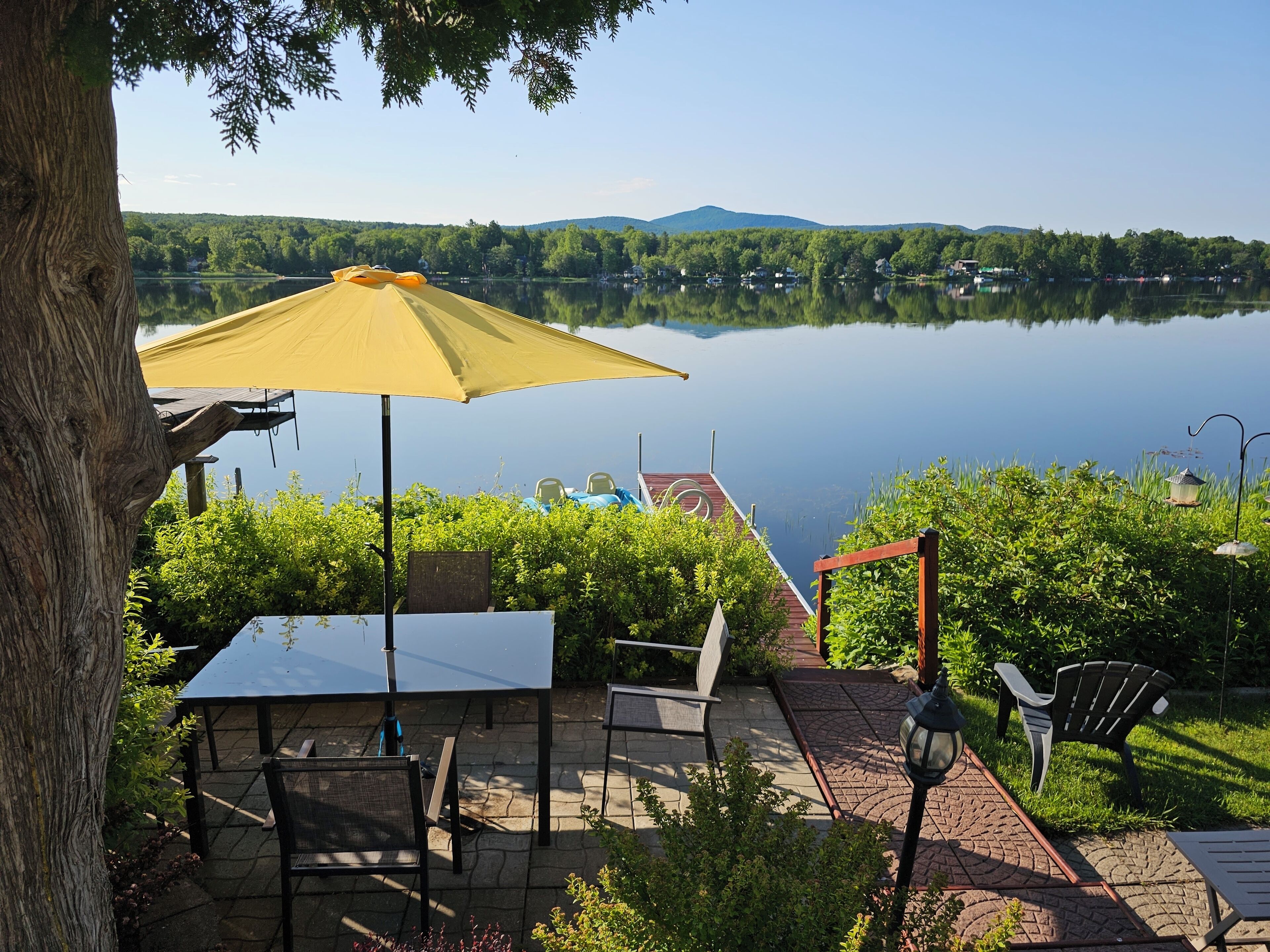 A lakeside patio with a table and chairs, a yellow umbrella, and a view of the lake and hills.