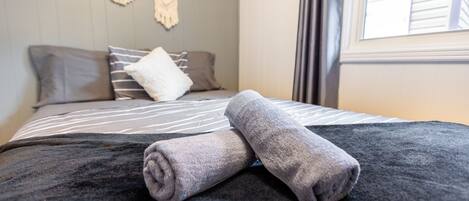 Cama bem arrumada com um cobertor escuro e duas toalhas enroladas, um travesseiro listrado cinza e branco, e um painel de macramê na parede.