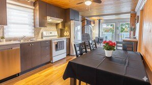 A modern kitchen with wooden ceiling, stainless steel appliances, and a dining area with a table set for two. - Cottage Tranquility family waterfront mountains (Dunham)