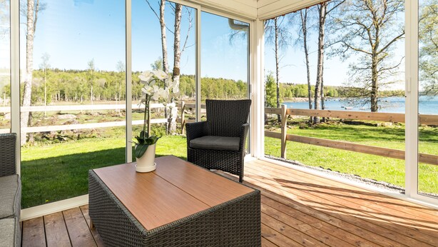 Un espacio al aire libre cubierto con piso de madera, mobiliario de mimbre y vistas a un lago y un bosque.