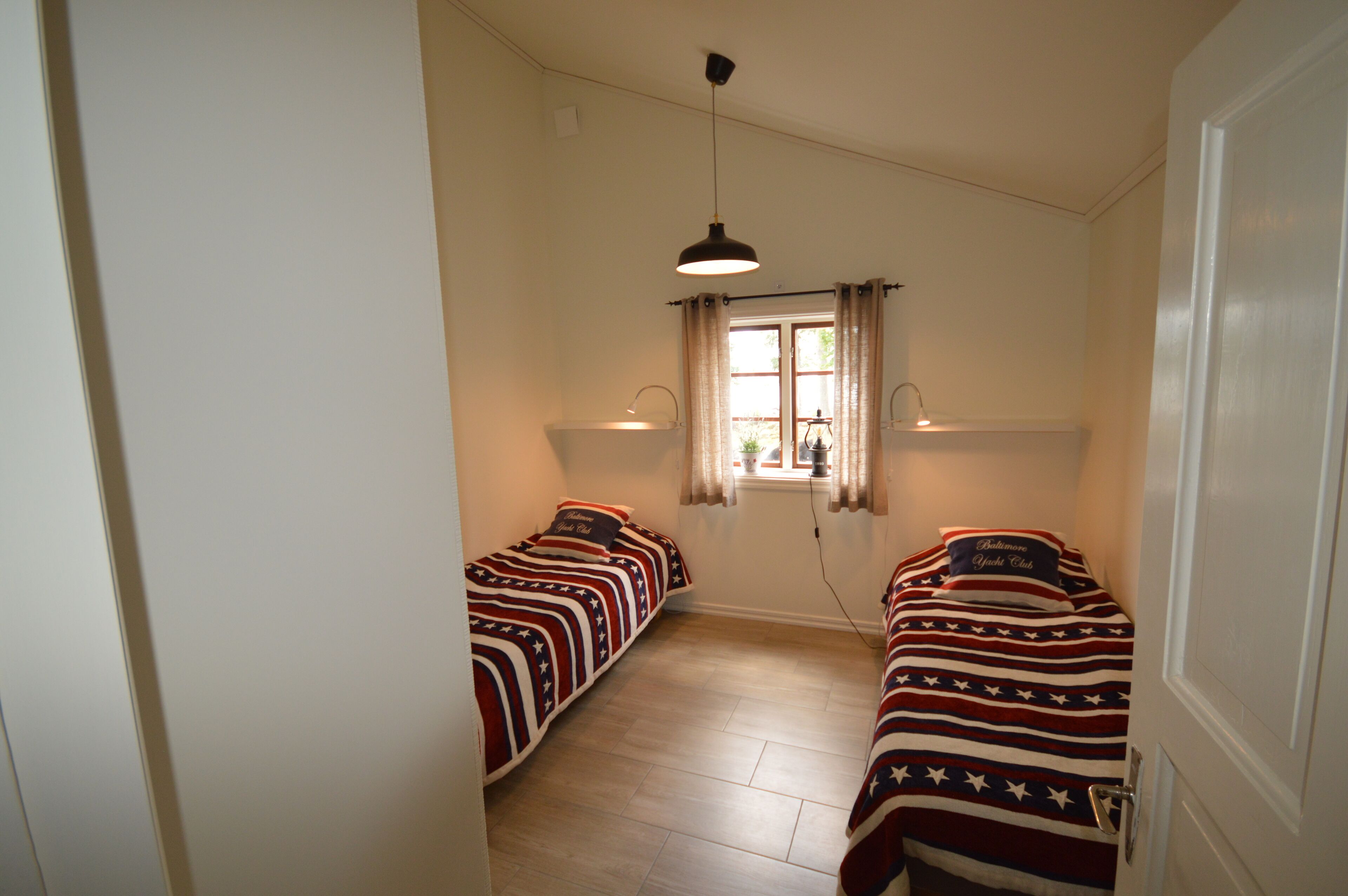 Et aflangt værelse med to senge, hver med et mønstret sengetæppe. Der er et vindue med gardiner, en hængende lampe og en dør til højre.