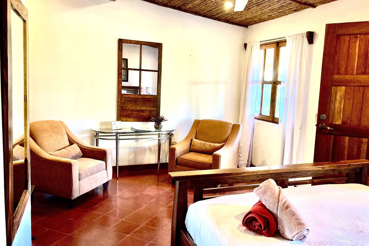 Una habitación de hotel con una cama, dos sillones, una mesita, un espejo y una puerta de madera.