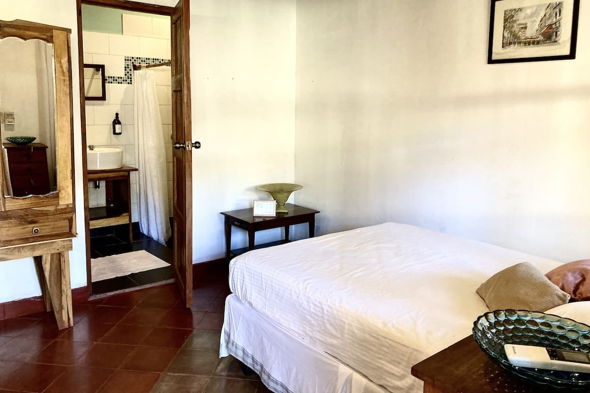 Un dormitorio con cama, mesita de noche, espejo, puerta que da al baño y un cuadro en la pared.