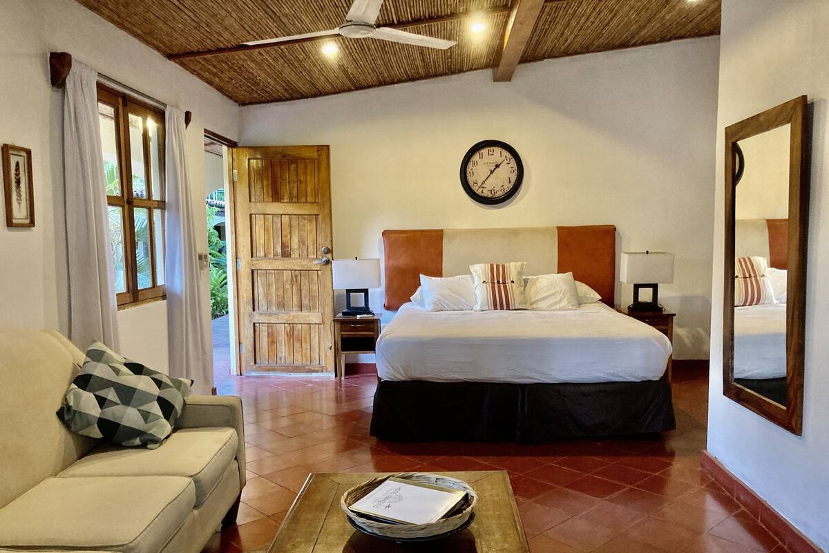 Habitación de hotel con cama, sofá, mesita de centro con bandeja, reloj y puerta que da al exterior.