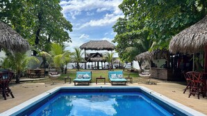 Un área de piscina con sillones de descanso, una estructura de techo de paja y vegetación tropical.