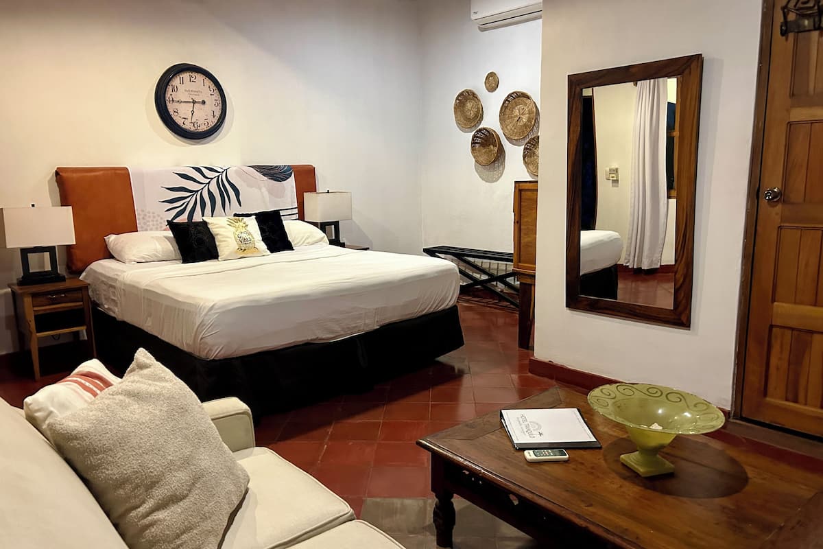 Una habitación de hotel con una cama, una mesita de noche, una lámpara, una mesa de centro de madera, un cuenco de vidrio verde, un espejo y un reloj de pared.