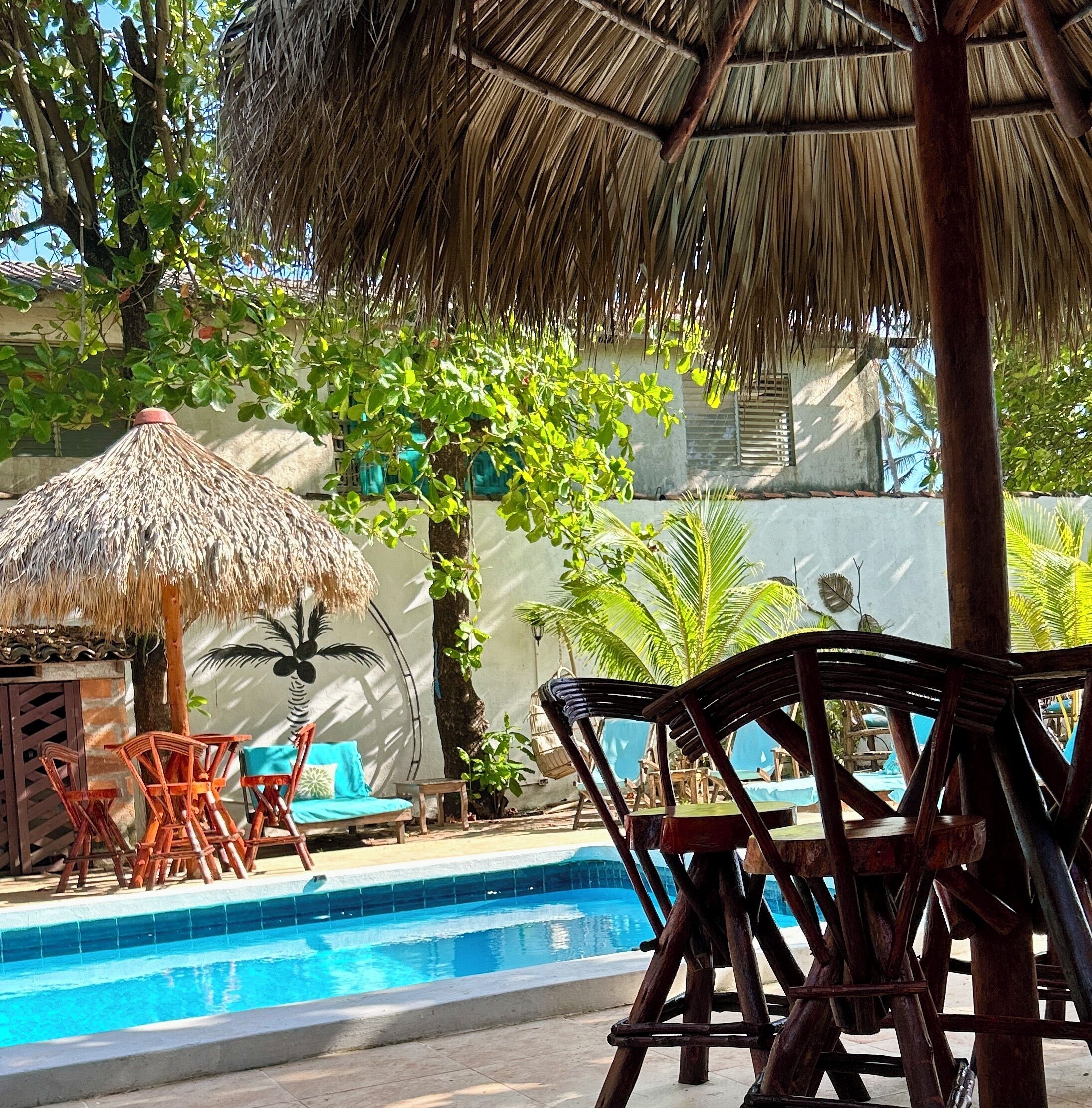 Un área junto a la piscina con una sombrilla de paja, sillas de madera y una palmera.