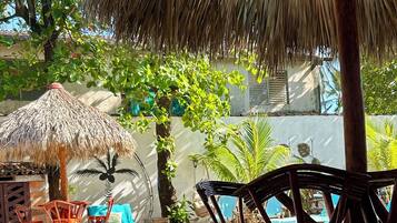 Un área junto a la piscina con una sombrilla de paja, sillas de madera y una palmera.