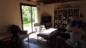 A living room with a sofa, armchair, and a television. There is a bookshelf and a window with a view of greenery. - Le Cottage, Country house 15 minutes from the beaches of Deauville (Saint-Julien-sur-Calonne)