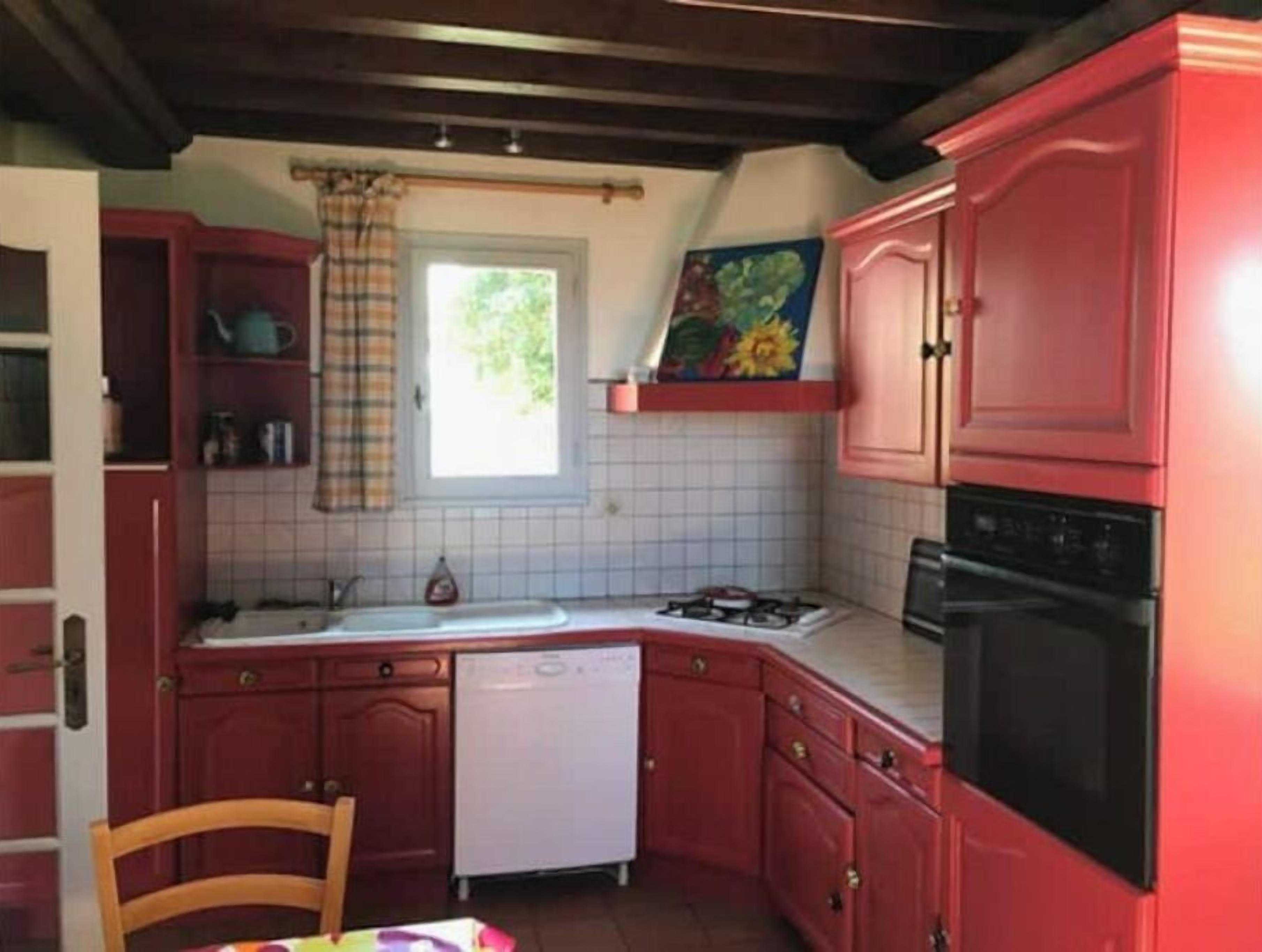 A compact kitchen with red cabinets, a white dishwasher, a stove, and a window with a curtain.