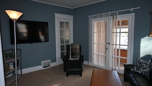 Ein Wohnzimmer mit Flachbildfernseher, einem schwarzen Ledersofa, einem Holztisch, einem karierten Sessel und einem Fenster mit weißen Rahmen und Jalousien.