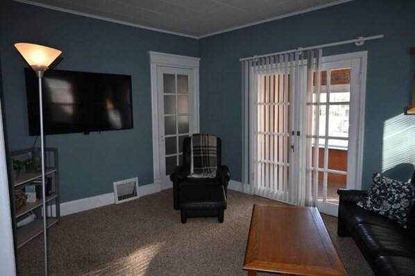 A living room with a flat-screen TV, a black leather sofa, a wooden coffee table, a plaid chair, and a white-framed window with blinds. - Lower flat steps from the lake and downtown (Port Washington)