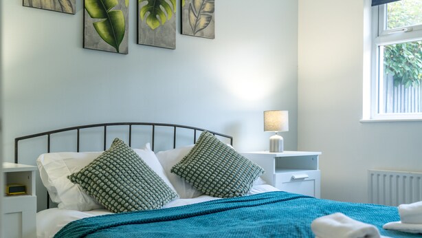 A neatly made bed with a teal blanket and two patterned pillows, a bedside lamp, and artwork on the wall. - Crawley Pet Friendly 1-Bedroom Apartment (Crawley)