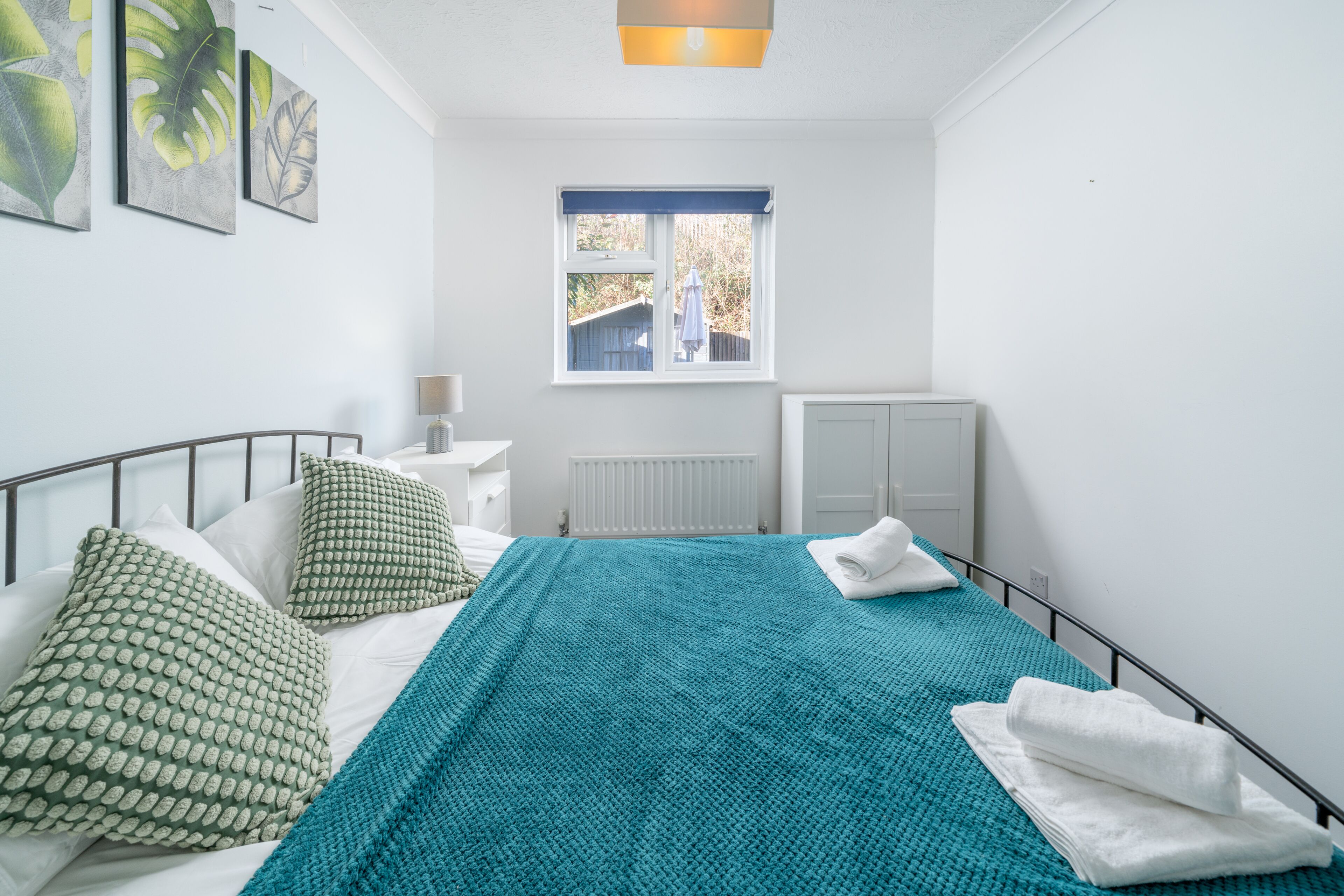 Una cama bien hecha con una manta color turquesa, almohadas blancas y una toalla blanca. Hay una ventana con vistas al exterior, un radiador y una pared con cuadros.