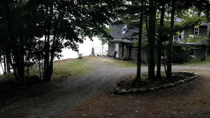 A gravel path leading to a rustic building with a porch, surrounded by trees and a body of water.